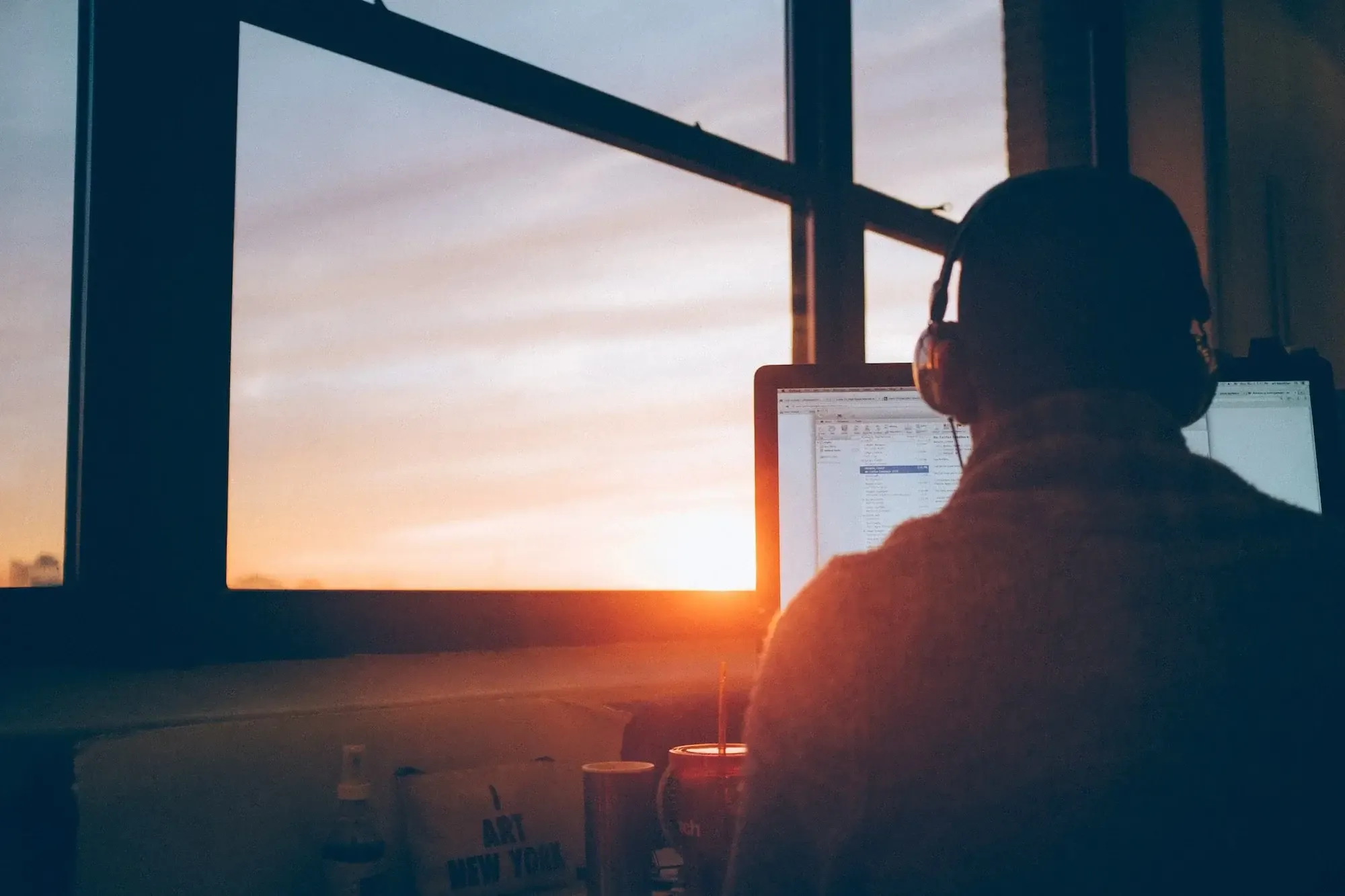 Remote worker seen from the back sitting at his desk by the window, sun goes down