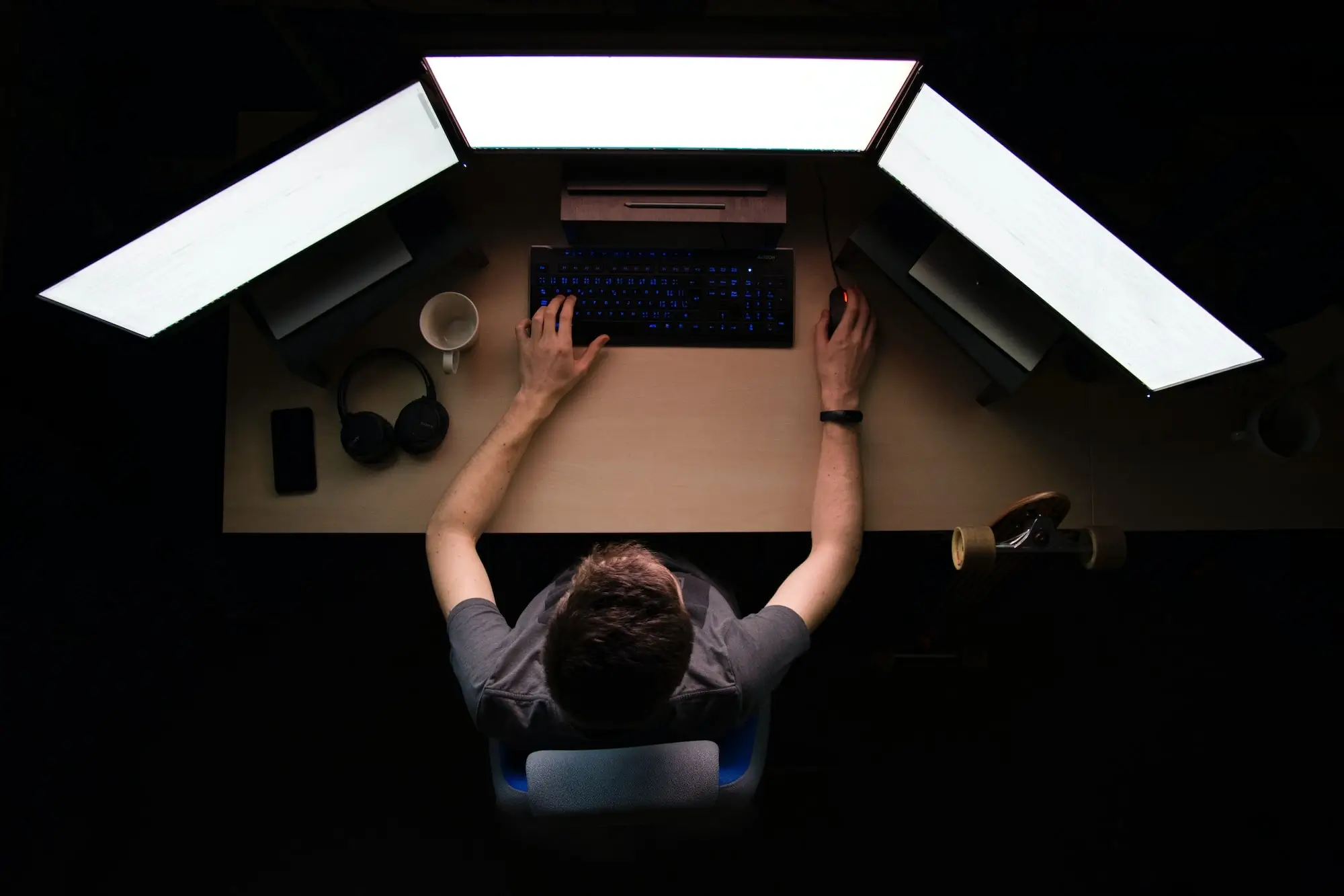 Software Developer sitting at his desk, three bright screens in front of him, a keyboard, coffee mug, smartphone and headphones laying next to him. Perspective from above