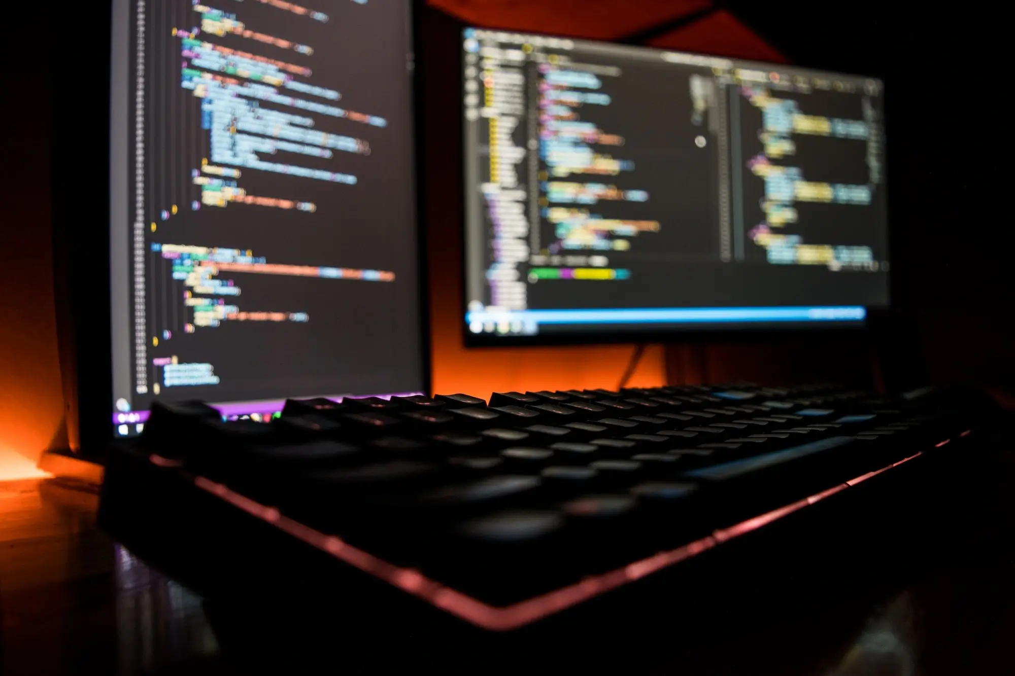 Tastatur und zwei Bildschirme mit Code auf einem Schreibtisch in einem dunklen Raum.