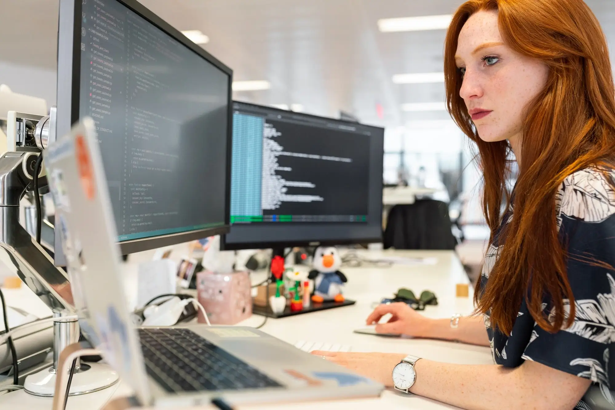 Eine Software-Ingenieurin sitzt an ihrem Schreibtisch mit zwei Bildschirmen und einem Laptop, arbeitet an Fehlern und kodiert ein Projekt
