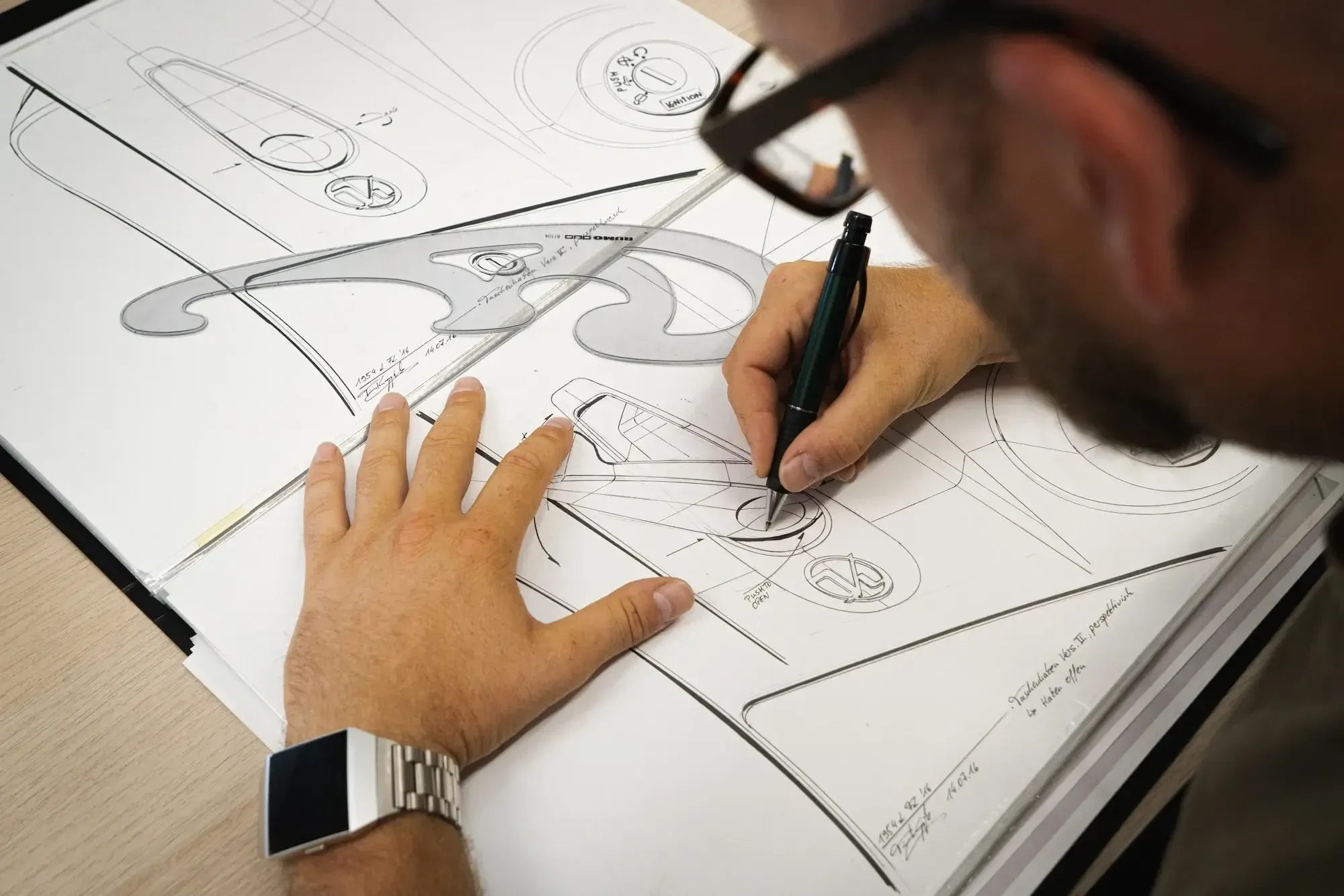 Mann mit silberner Uhr und schwarzer Brille, der Werkzeuge zum Skizzieren eines Produkts benutzt.