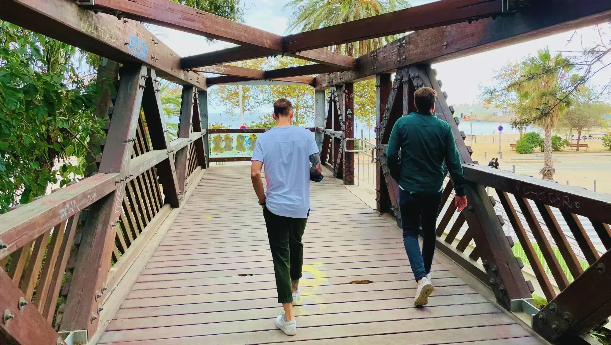 two men walking accross a wooden bridge in barcelona towards the beach, one wears a white shirt black pants, the other one wears a green shirt with jeans