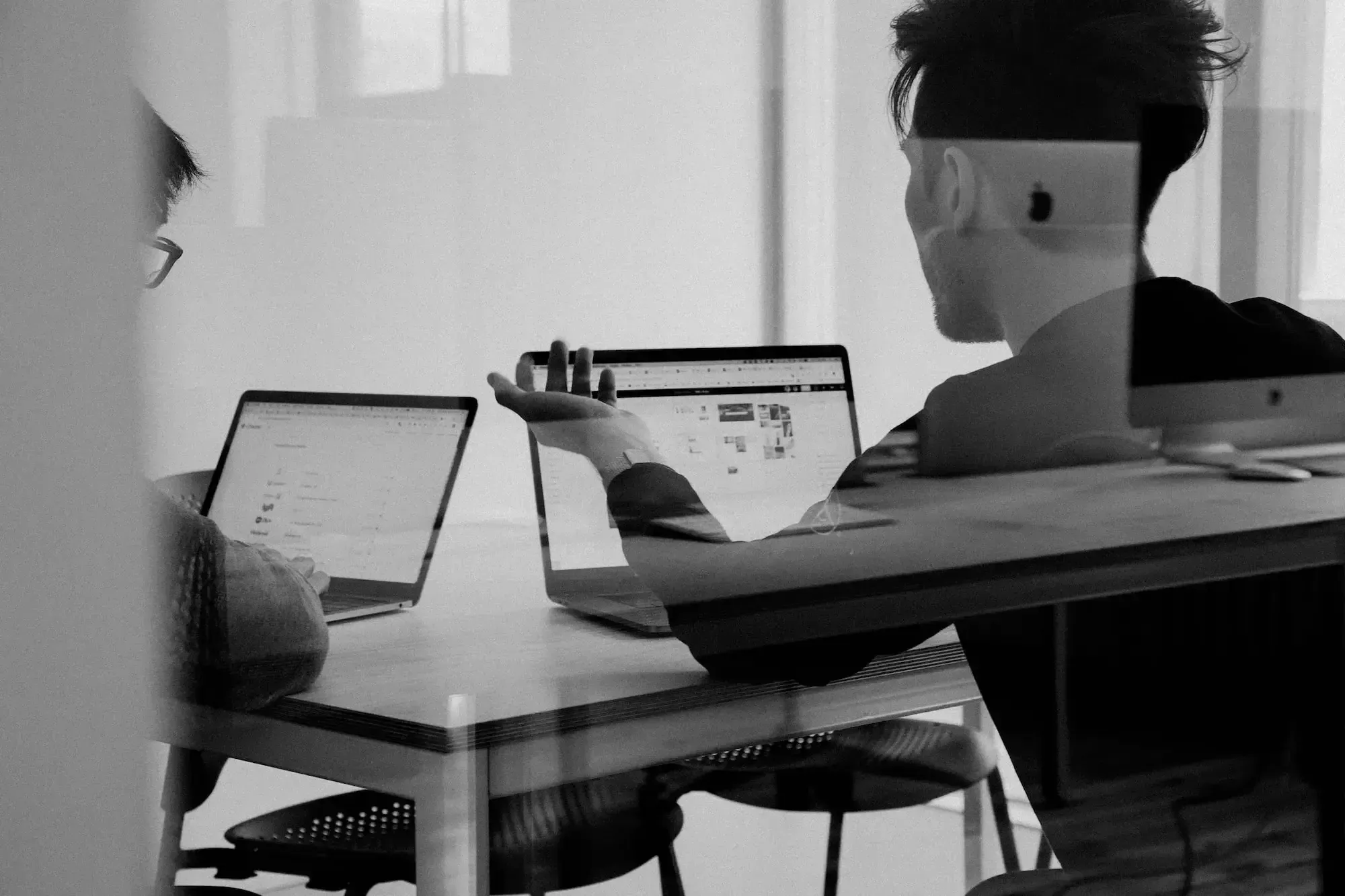 Two men are talking, laptops in front of them, black and white image.