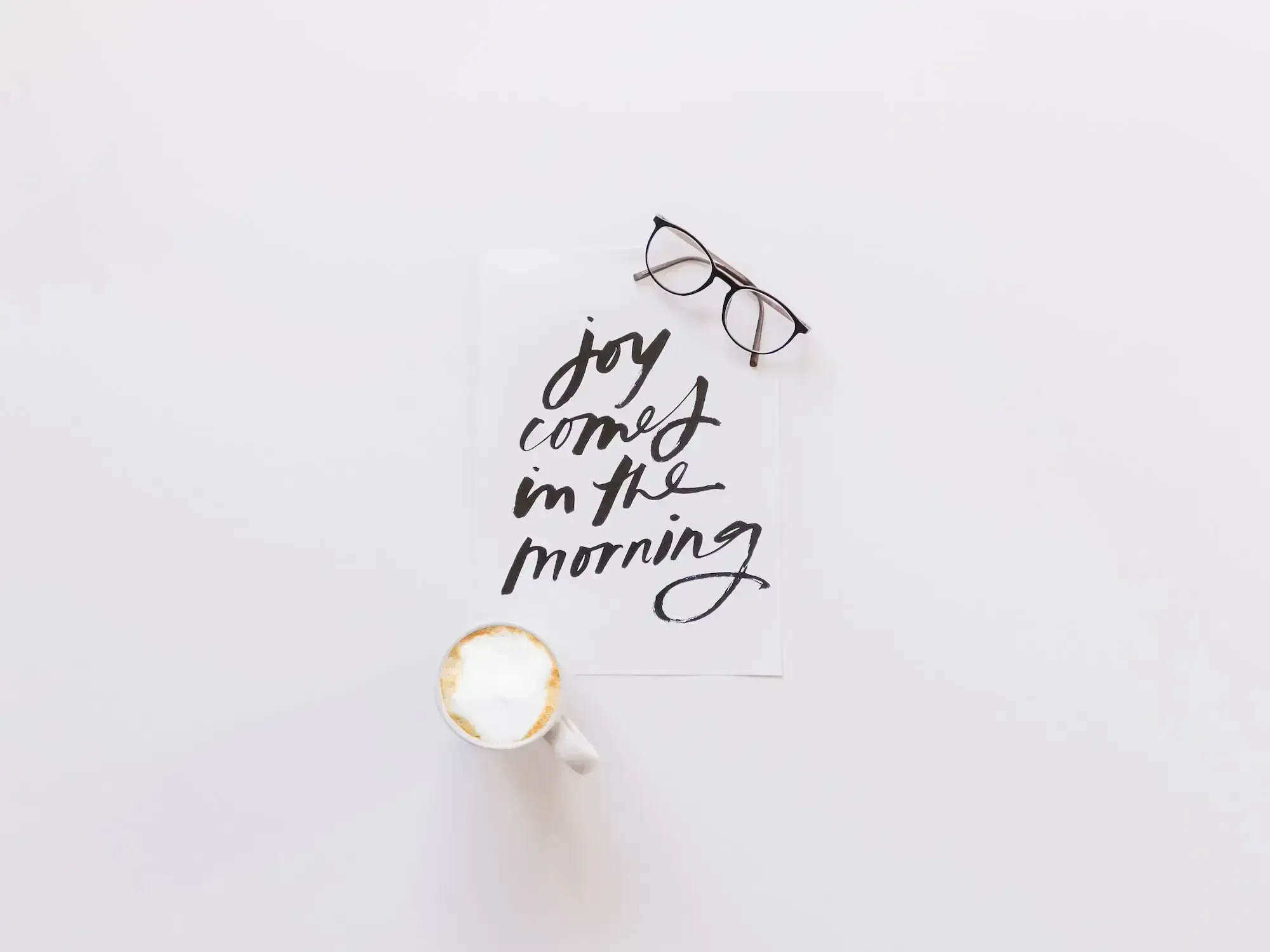 "joy comes in the morning" on a sheet of paper, glasses and a cup of cappuccino next to it.