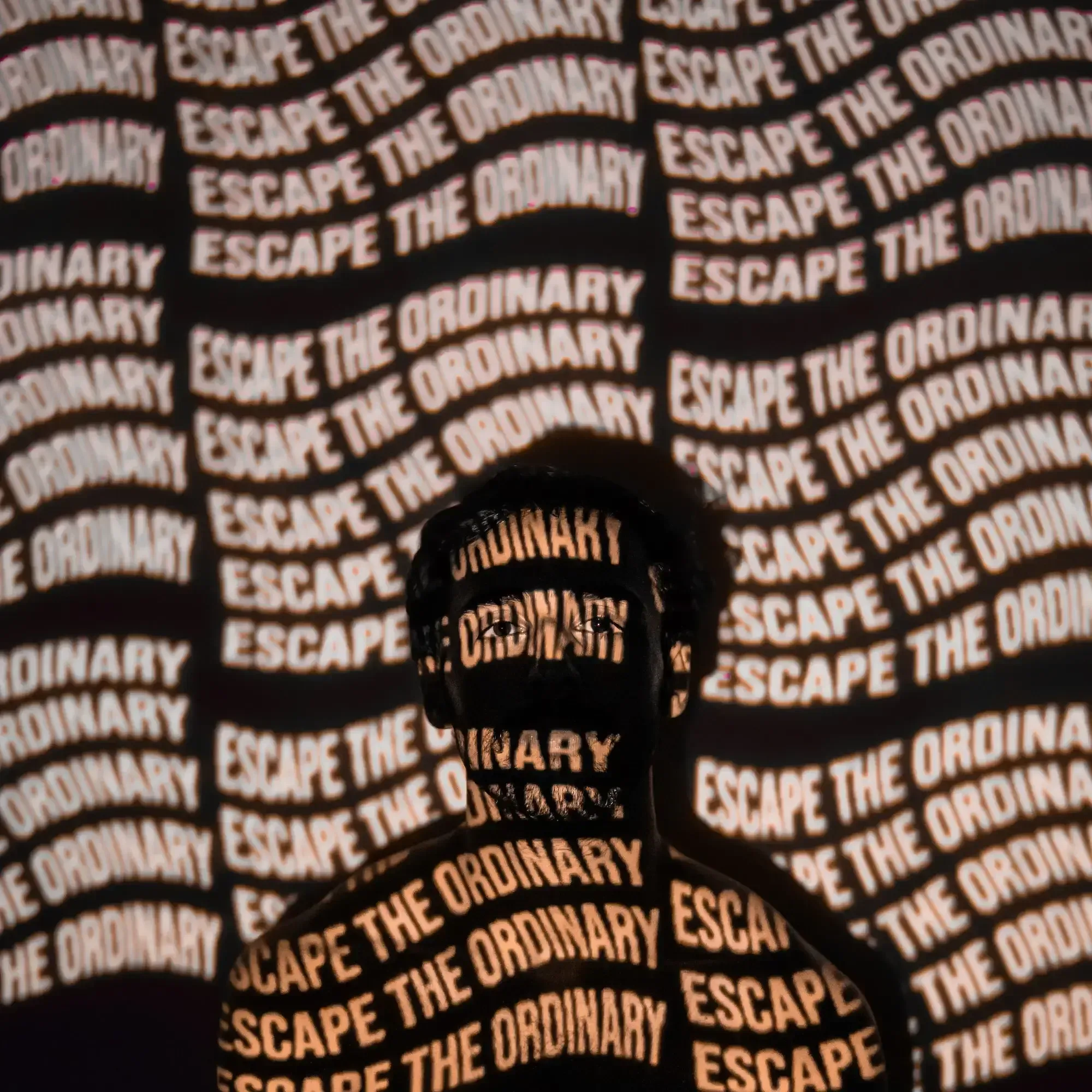 Man standing in front of a projector that throws yellow light at him, spelling "ESCAPE THE ORDINARY".