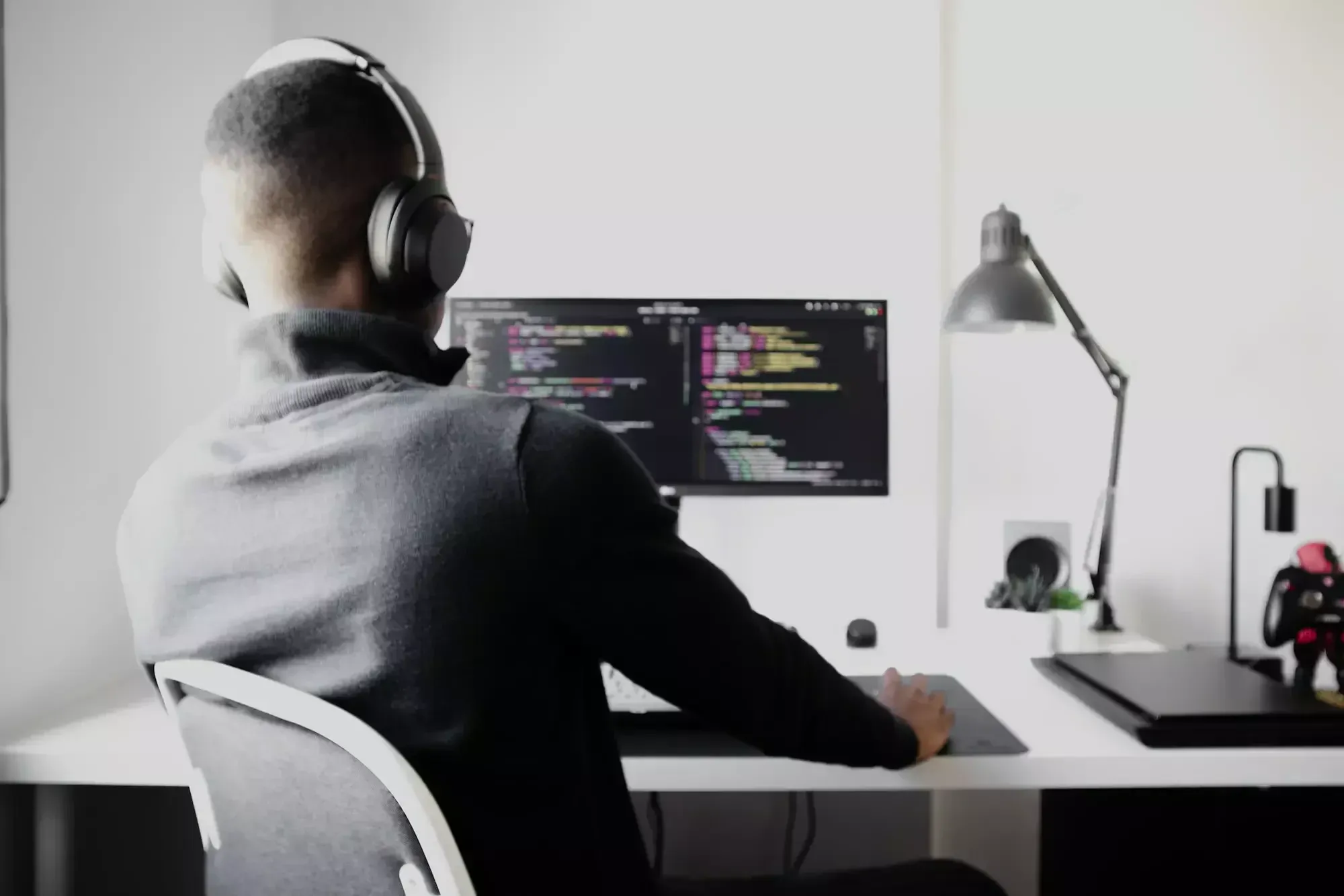 Mann in grauem Pullover und grauen Kopfhörern sitzt am Arbeitsplatz vor einem Bildschirm, auf dem ein langer Code zu sehen ist.