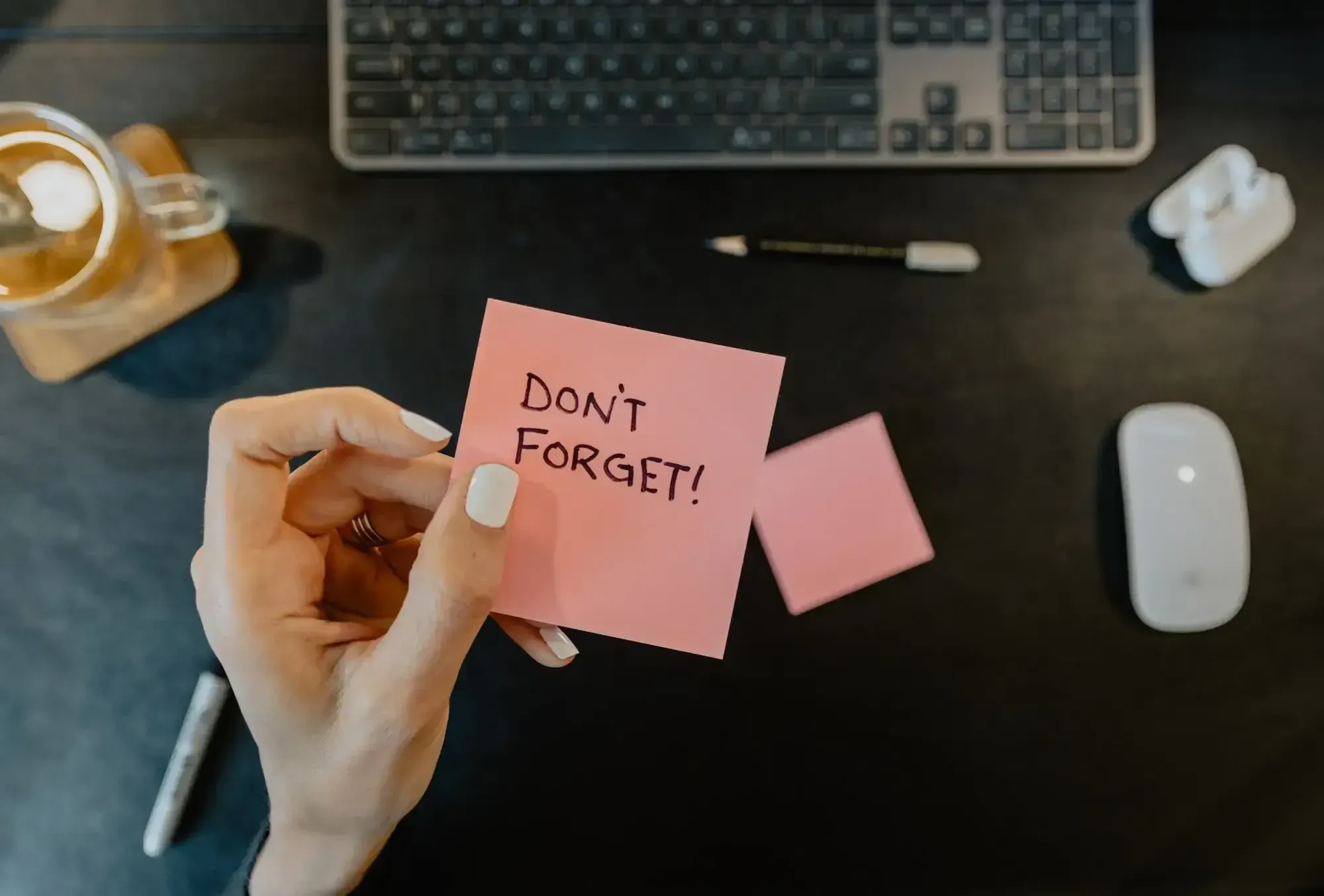 Hand with white nail polish and a golden ring holding a post-it with "Don't forget!" on it. In the background there's a desk with a keyboard, mouse, pens and a glas.