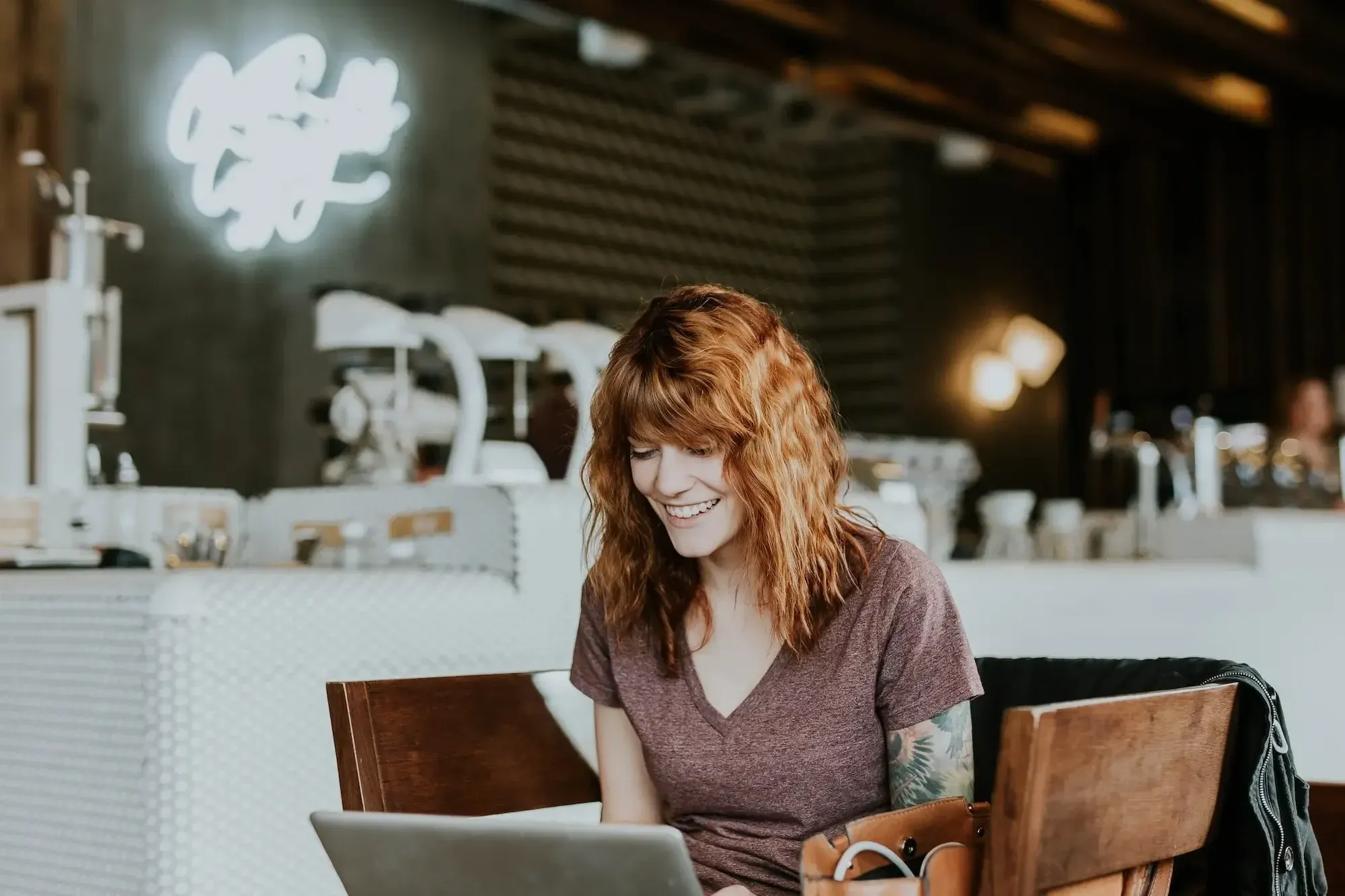 Eine lächelnde Frau mit roten Haaren und tätowiertem Arm sitzt in einem Café und hat ein virtuelles Meeting.