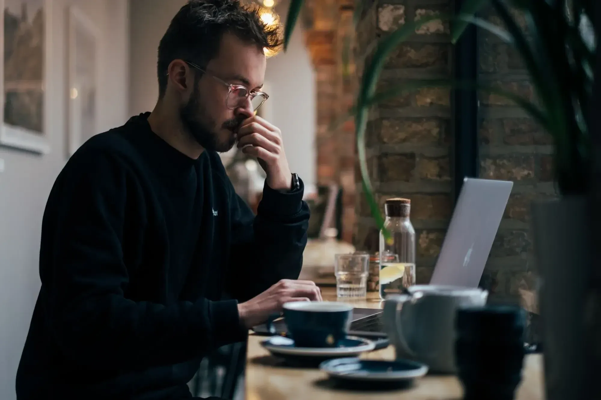 Man with black hoodie and glasses sitting in a brick wall cafe and working, cups standing next to him. 