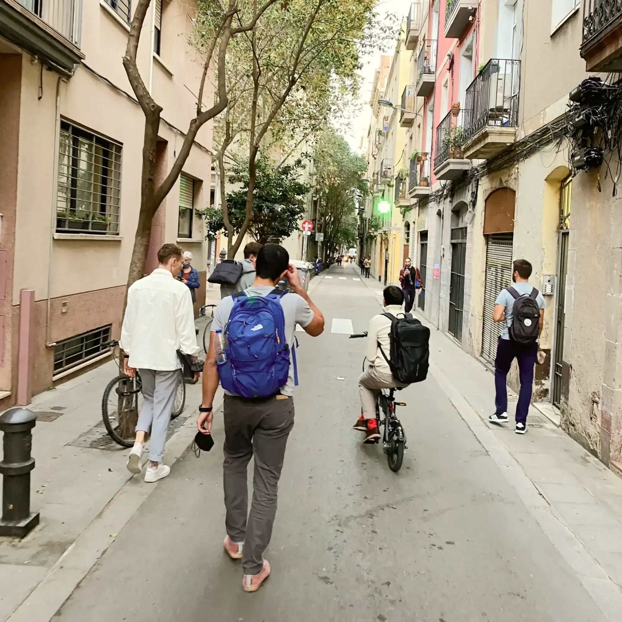 5 Men walking down a street in Barcelona, carrying backpacks, one is riding a bike