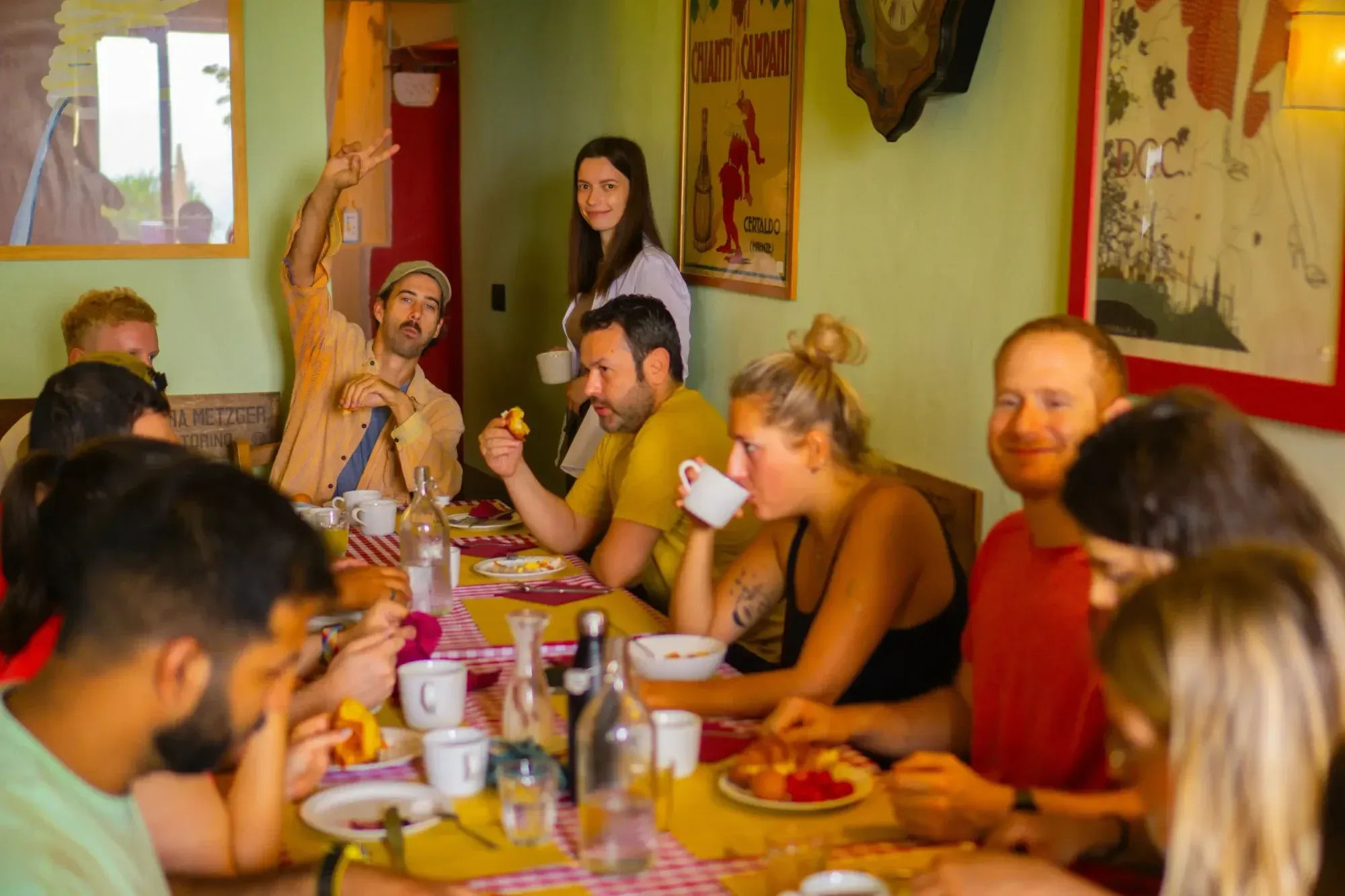 Ten people sitting on the breakfast table, eating and drinking. One man looking into the camera, raising his arm and showing a peace sign, woman to his right is holding a cup and smiling. 