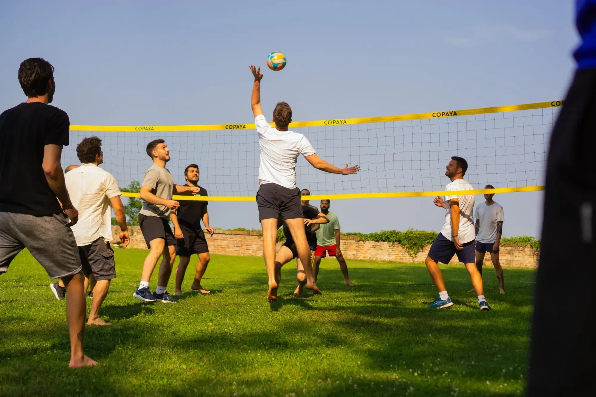Two groups of men playing volleyball. One man is jumping and hitting the ball right over the net. They are all wearing shorts and t-shirts, standing on grass in the sun.