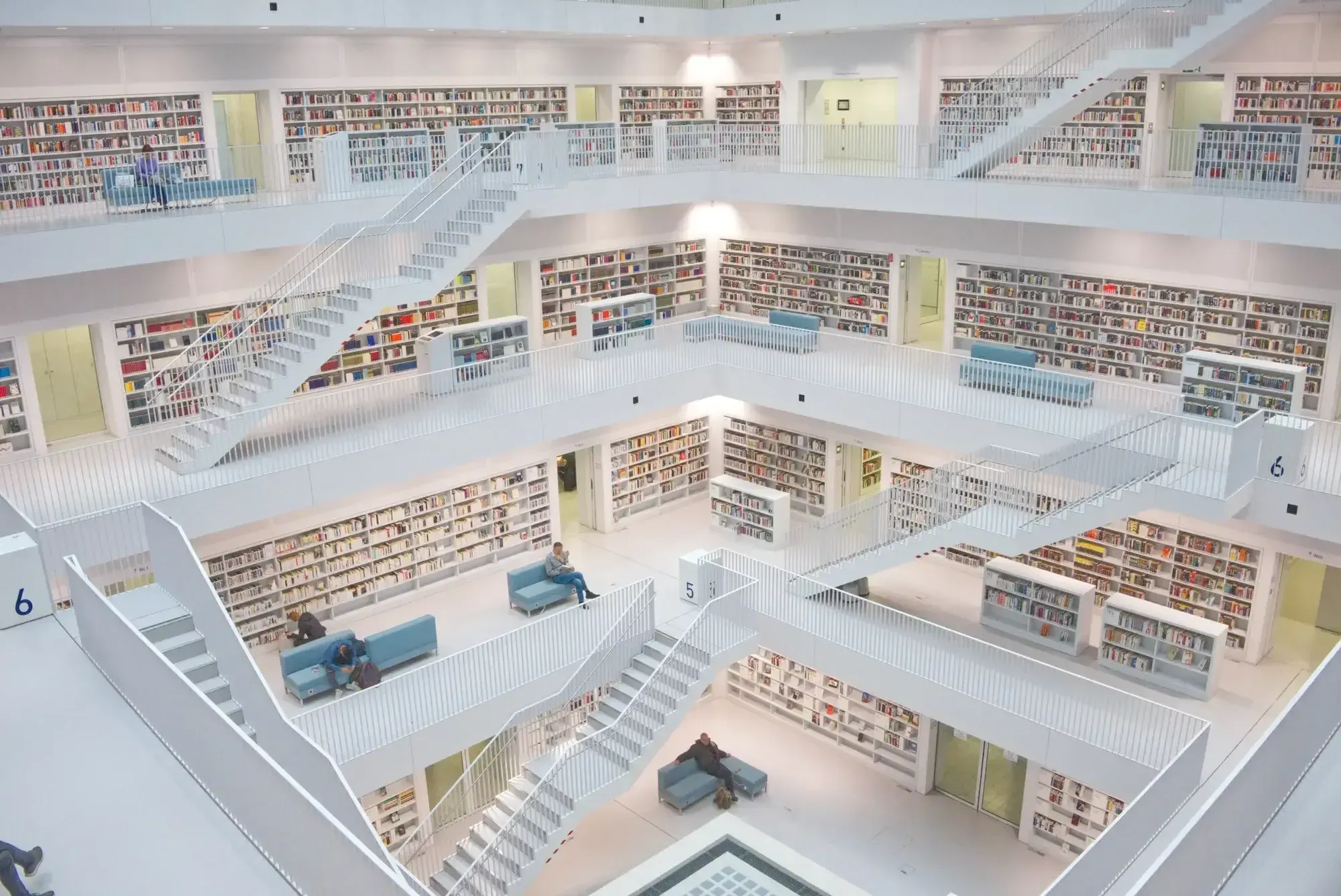 Huge library with white floors and white staircases and grey sofas, some people reading on them.