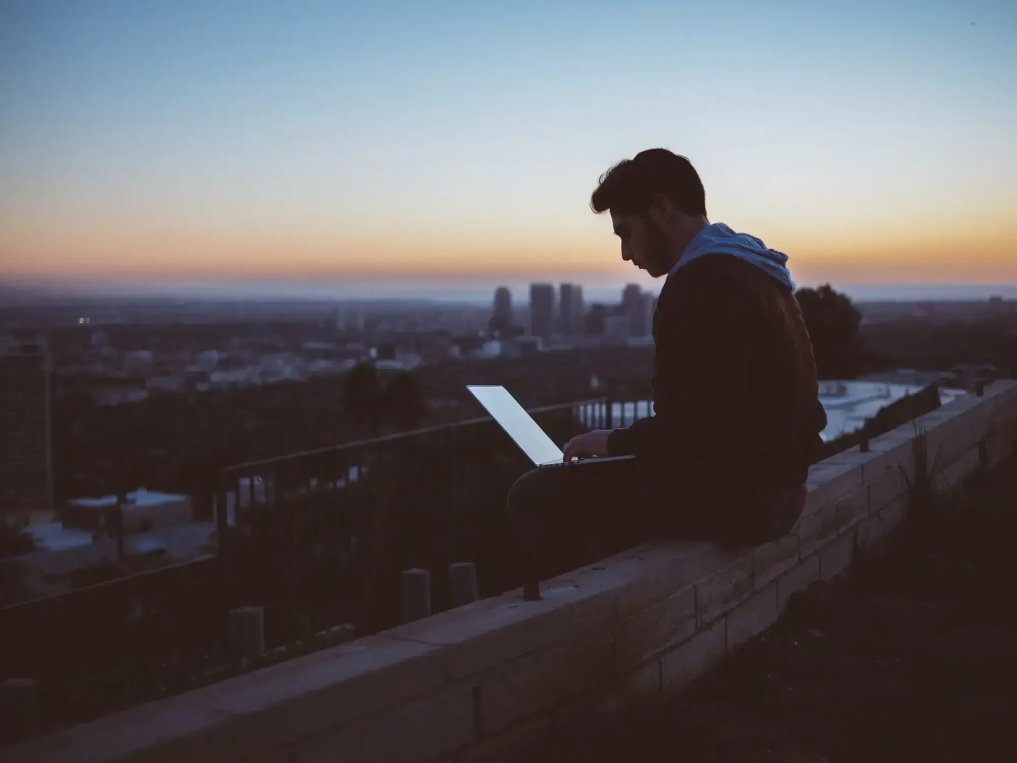 Man sitting on a roof terrace at sunset working on a laptop, city skyline in the back.