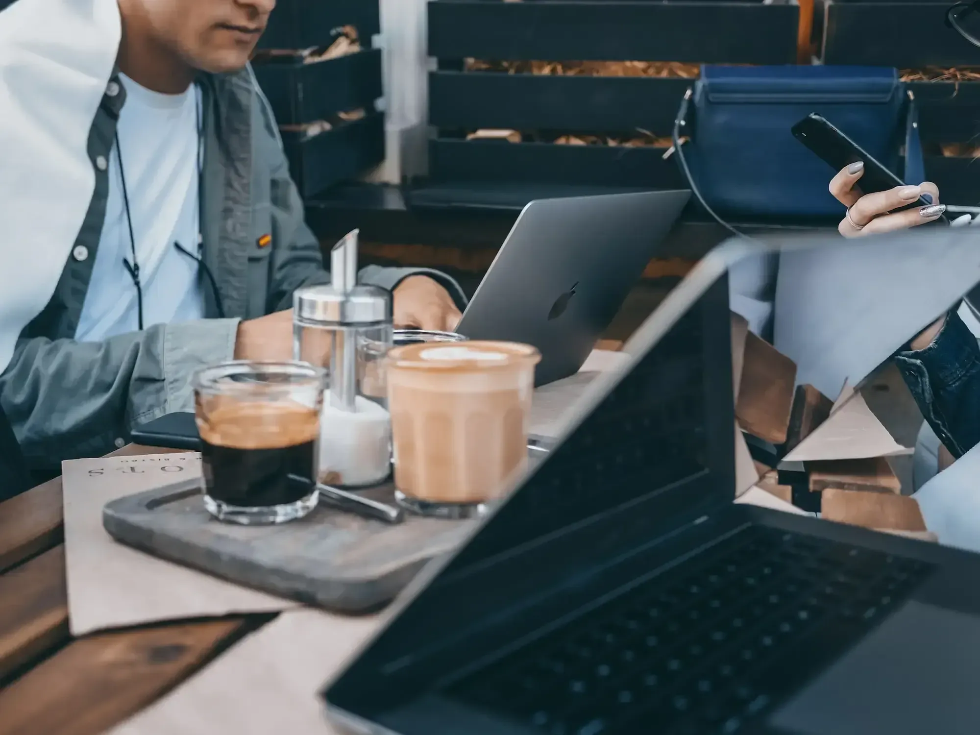 Drei Leute arbeiten draußen an ihren Laptops, zwei Tassen Kaffee und Zucker stehen auf dem Holztisch.