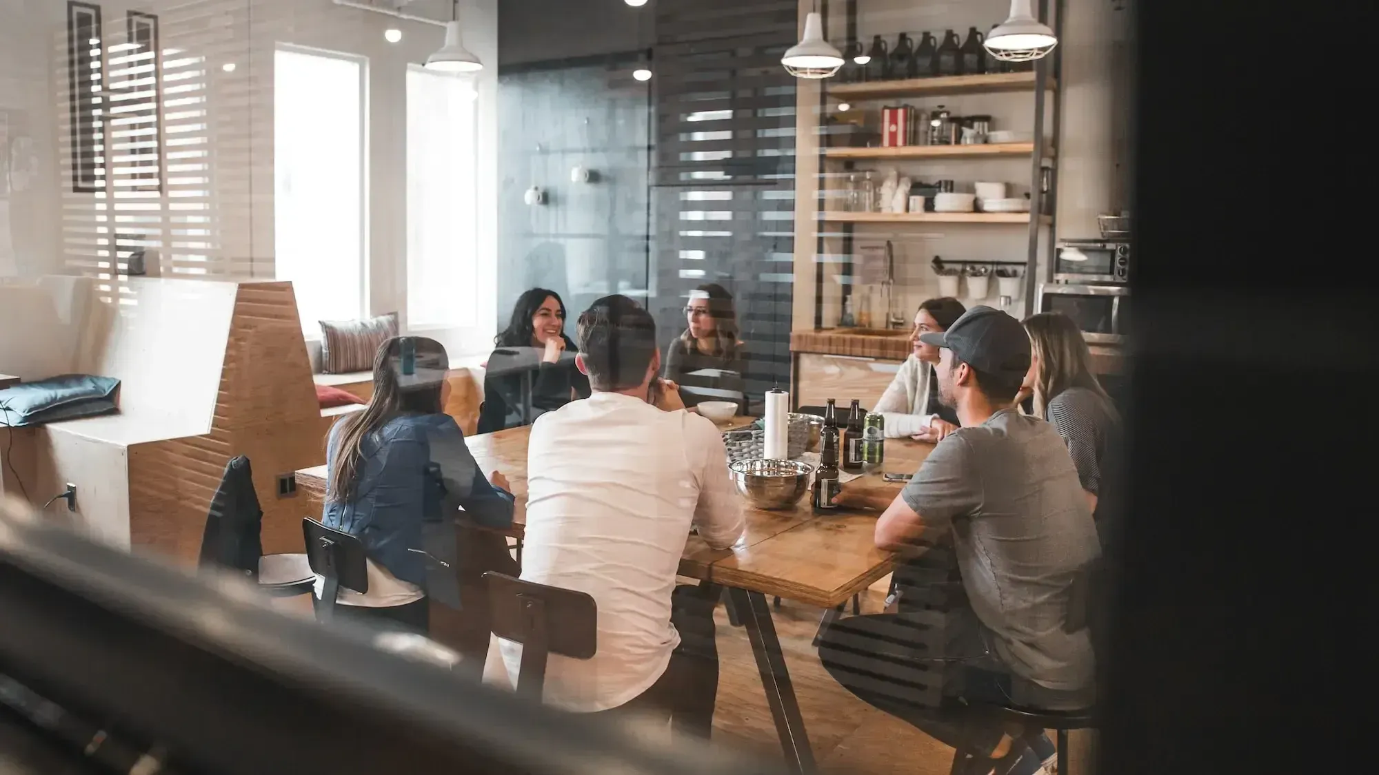 Sieben Leute sitzen an einem Holztisch in einem Meeting-Raum mit Glaswänden und einer Küche im Hintergrund.