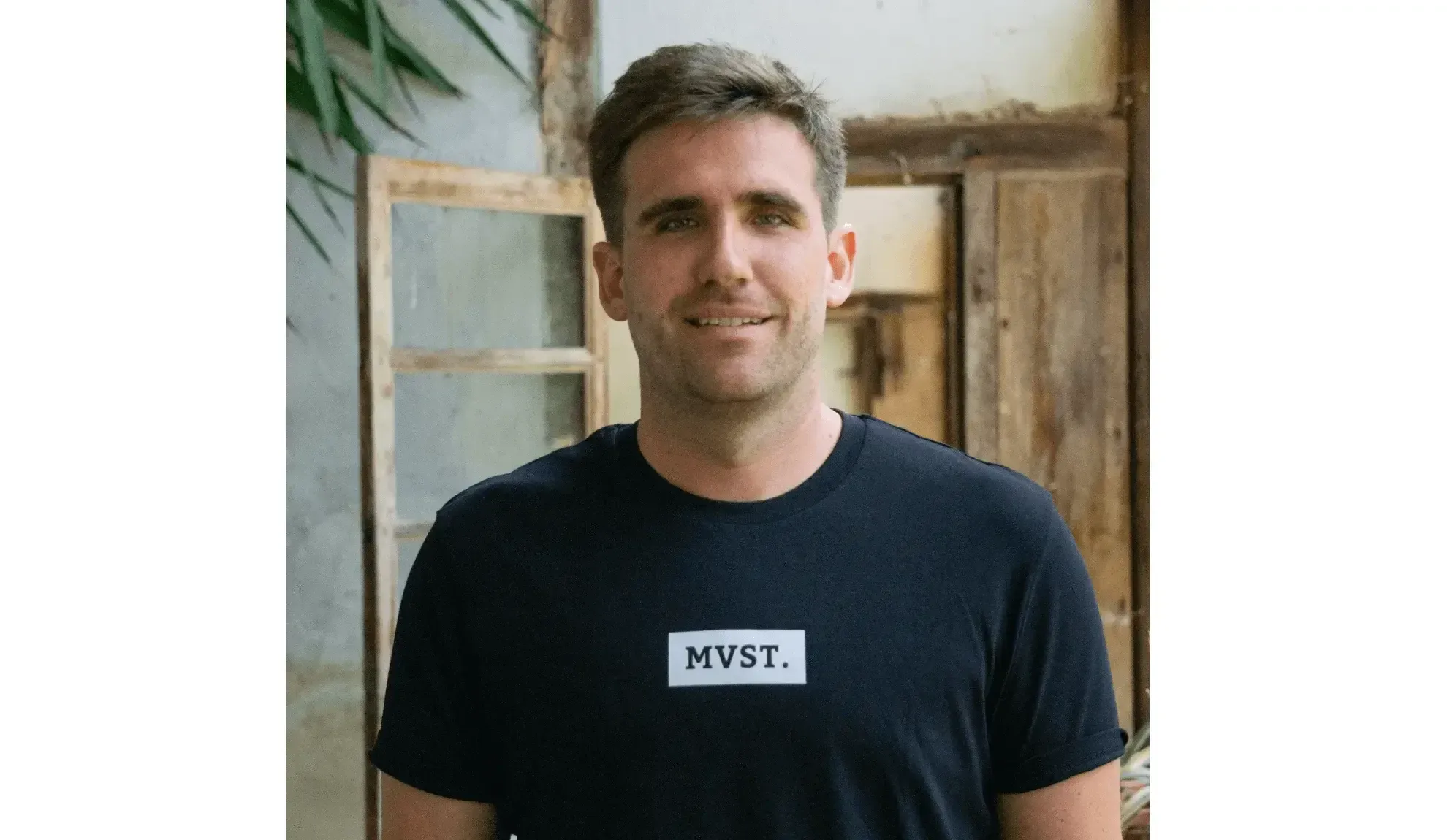 A Guy with dark blonde hair and a black tshirt standing in front of a wooden glass door. On the Tshirt is a Logo with the letters MVST