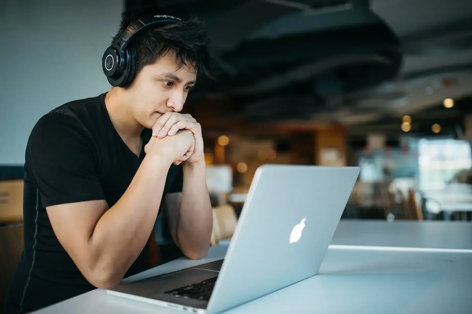 Man with a black shirt wearing headphones, looking at his laptop in front of him, concentrating with his hands on his chin.
