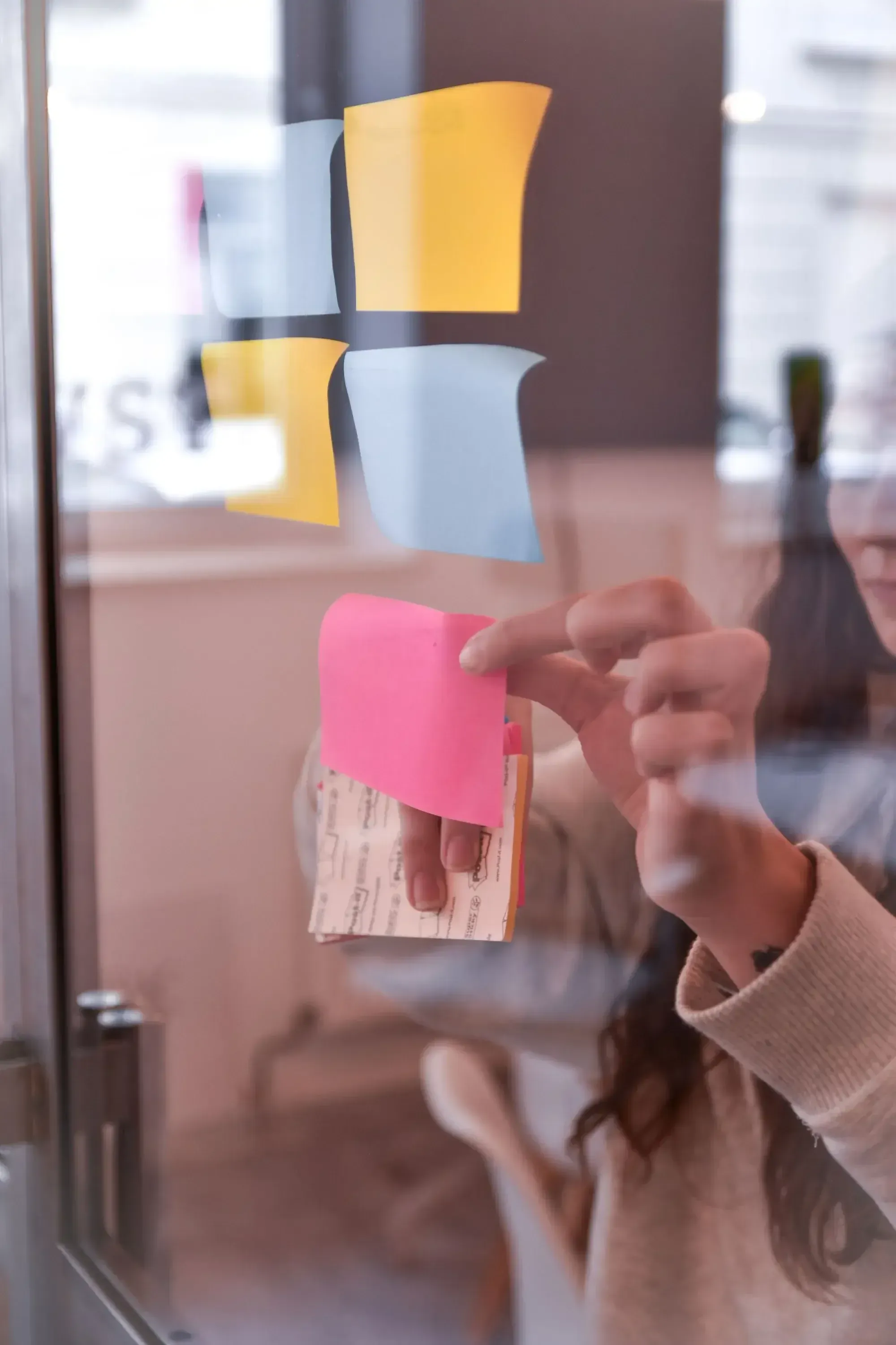 Woman with long brown hair and beige jumper sticks colourful post-its on a glass wall.