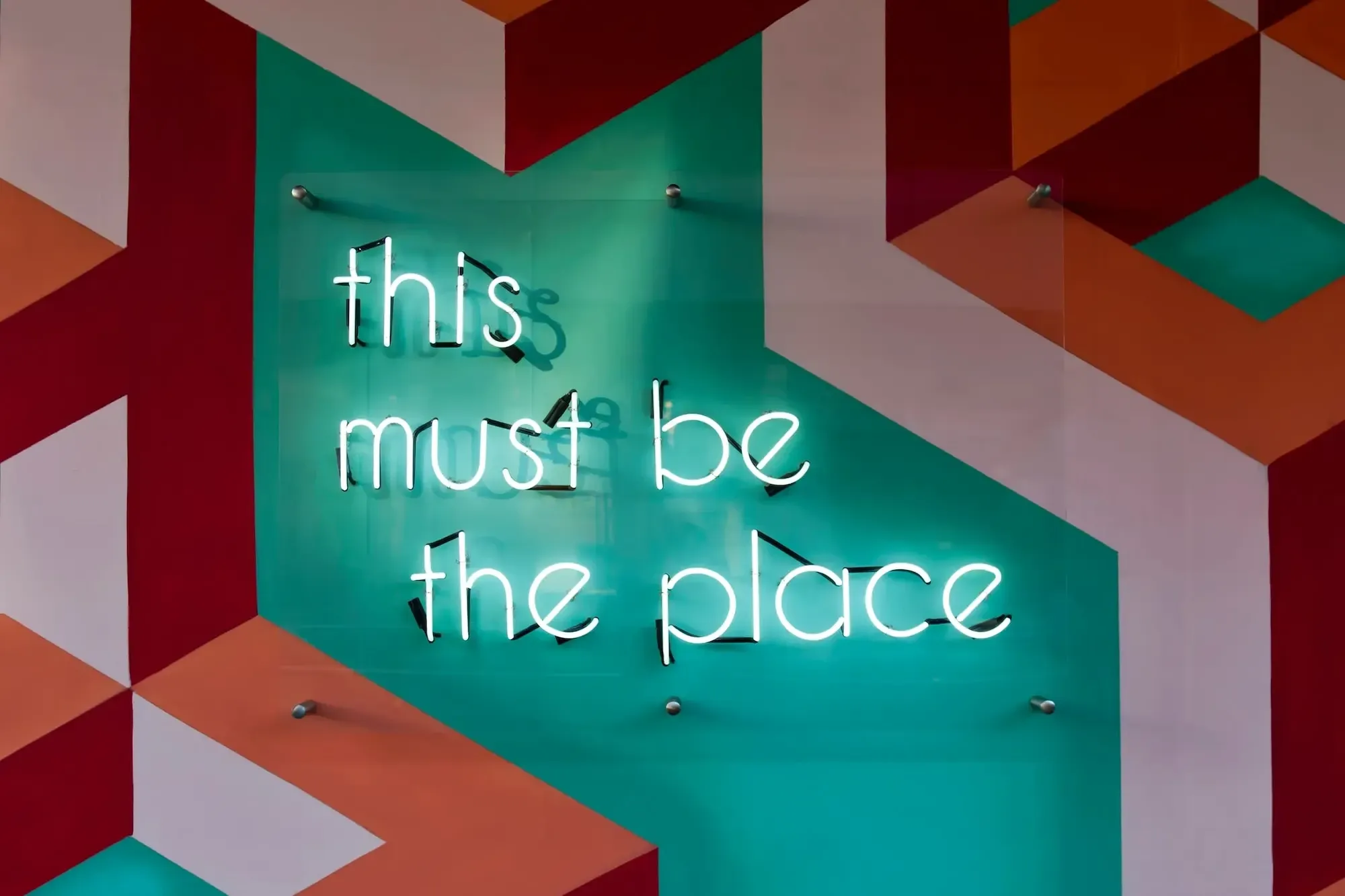 Pink, turquoise and red 3D painting, white neon lights in the middle spelling "this must be the place"