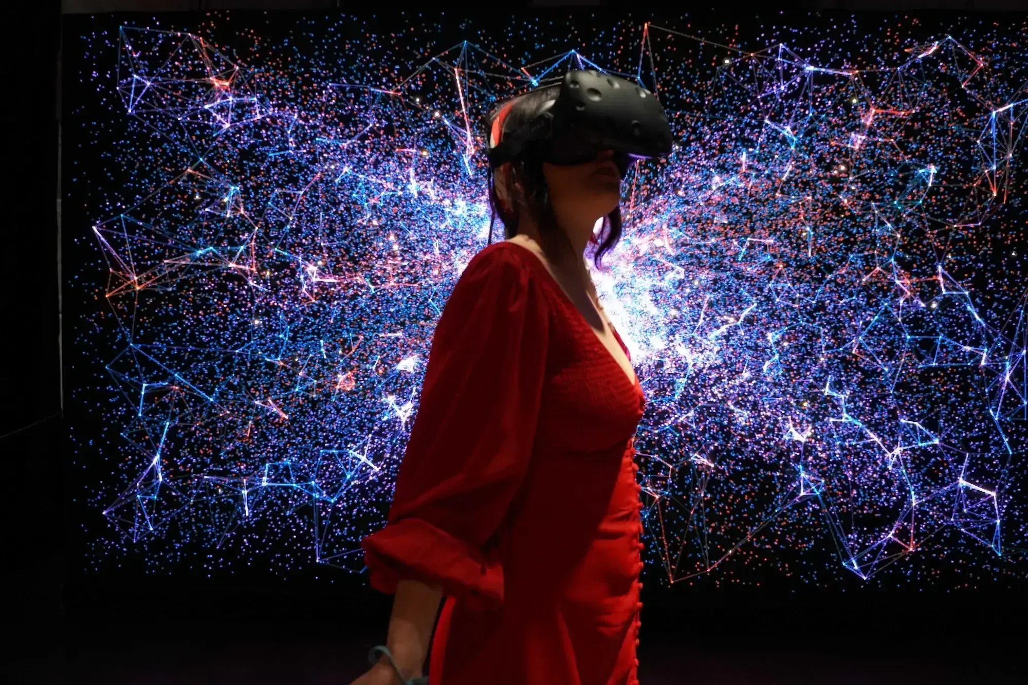 Frau in rotem Kleid mit VR Brille auf in einem dunklen Raum voller bunter Lichtpunkte.